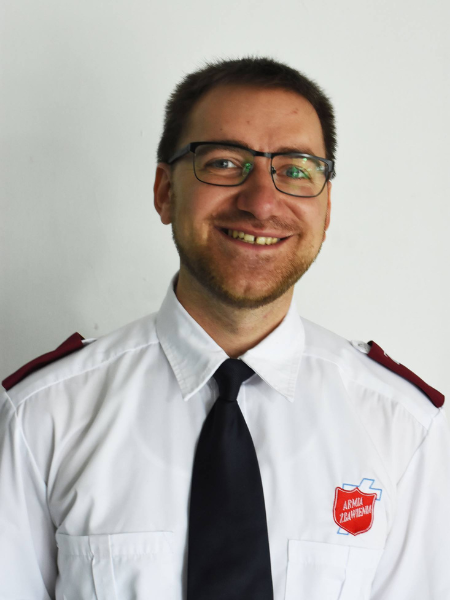 Portret ciemnowłosego mężczyzny w okularach. Ubrany w białą koszulę i krawat. Na lewej piersi czerwona tarcza Armii Zbawienia.