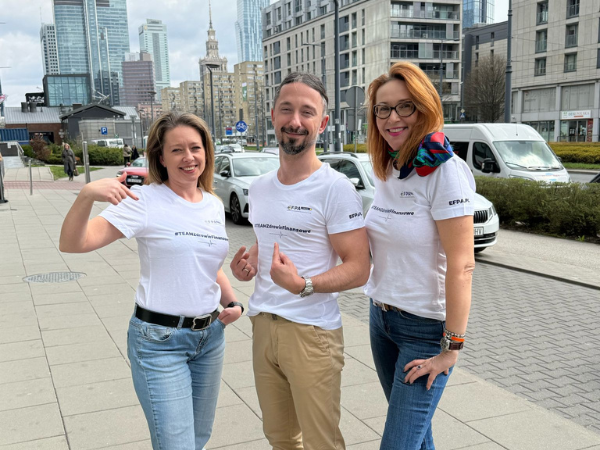 Dwie kobiety i mężczyzna w białych koszulkach, stojący na chodniku na tle pałacu kultury