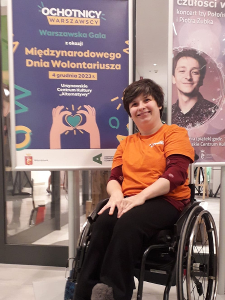 Kobieta na wózku inwalidzkim w pomarańczowej koszulce z napisem Fundacja Avalon przed plakatem z napisem "Warszawska Gala z okazji Międzynarodowego Dnia Wolontariusza" 