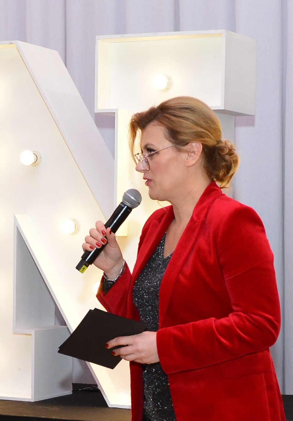 Kobieta w spiętych rudych włosach i okularach ustawiona bokiem do kamery. Jest ubrana w czerwony żakiet i błyszczącą czarną bluzkę. W prawej ręce trzyma mikrofon, a w lewej plik czarnych kartek.