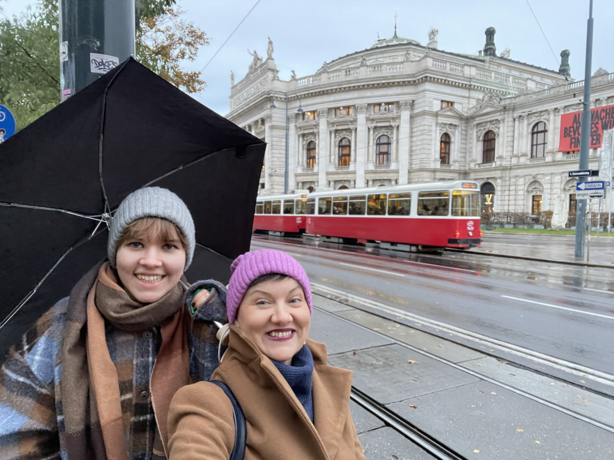 Dwie kobiety stojące po lewej stronie zdjęcie na tle kremowego budynku i czerwonego tramwaju. Jedna kobieta ma krótkie blond wlosy, szarą czapkę oraz płaszcz w niebiesko-brązową kratkę. W lewej ręce trzyma czarny parasol . Druga z kobiet ma krótkie czarne włosy, fiołkową czapkę oraz brązowy płaszcz z postawionym kołnierzem. 