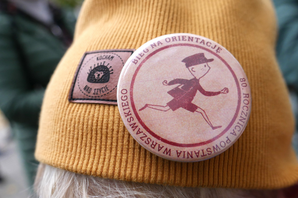 Żółta czapka z przypinką z napisem "Bieg na orientację. 80. rocznica Powstania Warszawskiego"