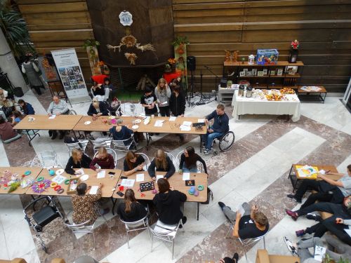Zdjęcie wykonane z lotu ptaka: obraz z warsztatów międzypokoleniowych, na zdjęciu stoły, oraz młodzież i seniorzy wykonujący rękodzieło