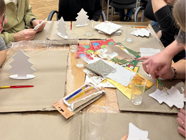 Jasnobrązowy stół zastawiony przyborami plastycznymi. Na stole ułożone są serwetki, modele choinek, farby, kubeczki z wodą oraz pędzle.