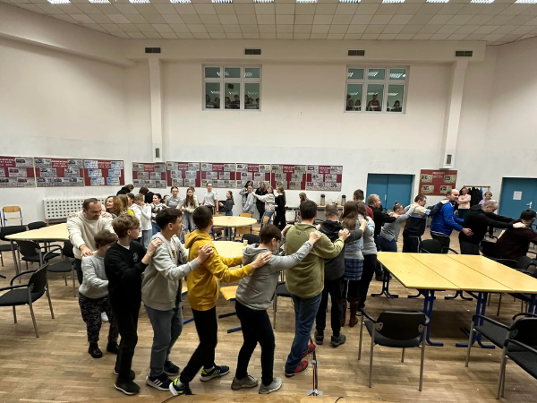 Grupa ludzi w różnym wieku na sali gimnastycznej stojąca jeden za drugim i trzymająca ramiona osoby stojącej przed nią.