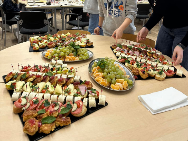 Jasnobrązowy stół zastawiony małymi przekąskami, na przykład roladkami, kanapkami oraz owocami.