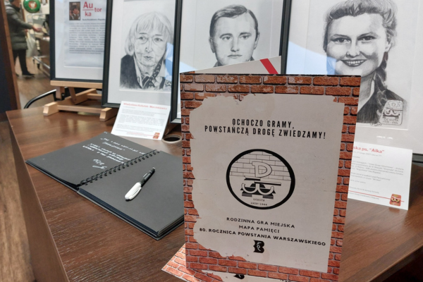 Stół, na którym rozłożone są ulotki dotyczące organizowanej gry terenowej. Na dalszym planie znajdują się portrety trzech