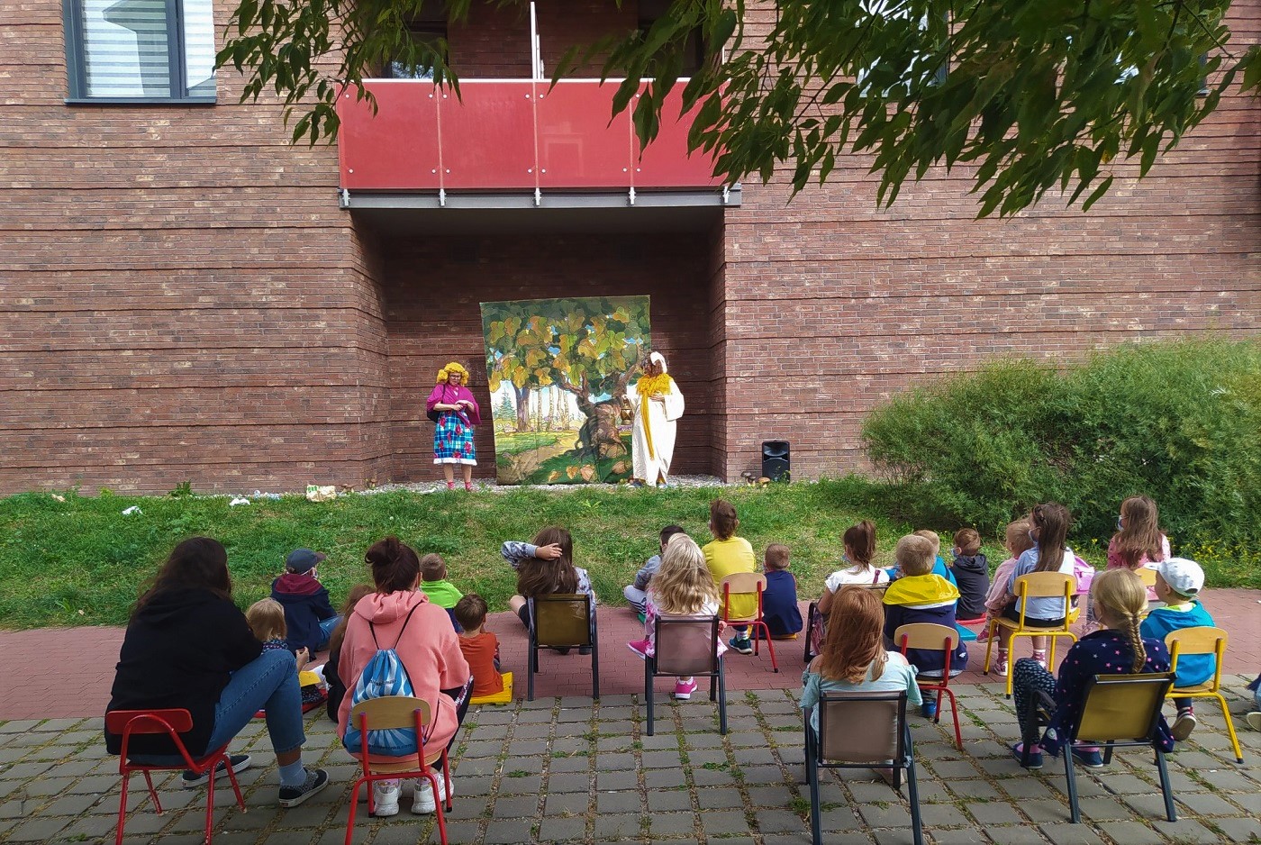 Przedstawienie przed biblioteką, dwóch performerów oraz widownia na krzesłach