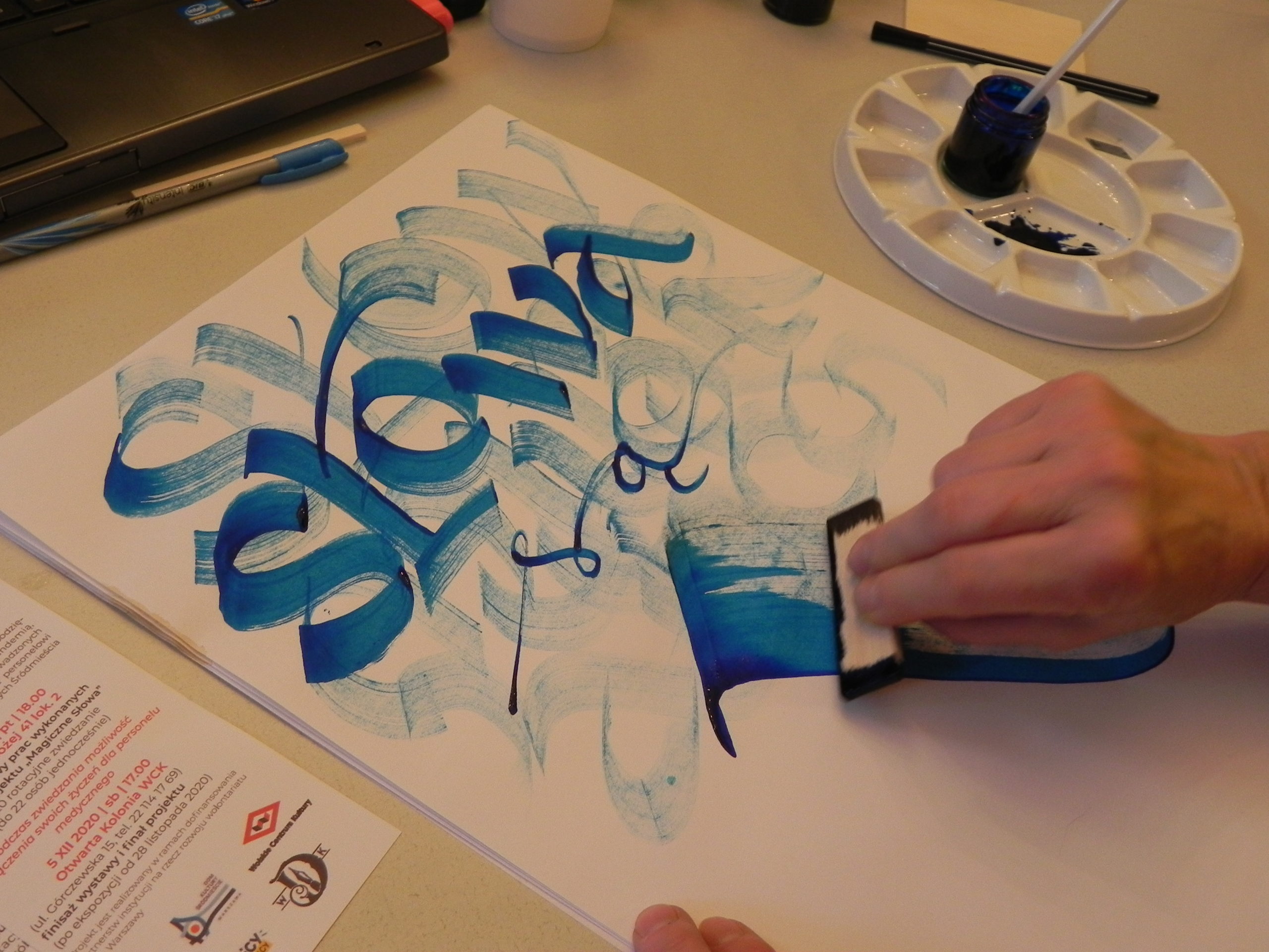 Zdjęcie z warsztatów kaligrafii przedstawiające w przybliżeniu proces tworzenia kaligrafii - zbliżenie na kartkę papieru niebieskim napisem "słowa", tworzonym krawędzią sześciennego przedmiotu zanurzonego w farbie