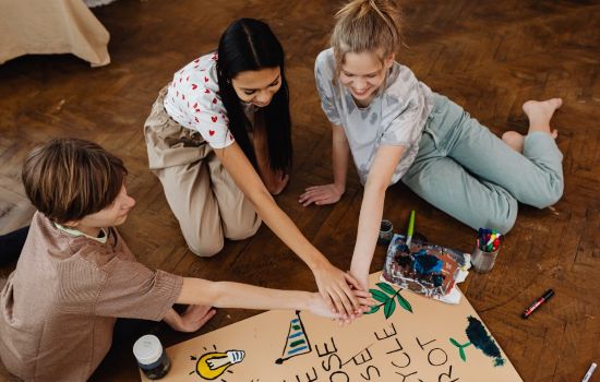 Grupa trójki młodych ludzi siedzących na podłodze przy stworzonym przez siebie plakacie, wyciągają przed siebie prawą rękę, kładąc dłonie jedna na drugiej 