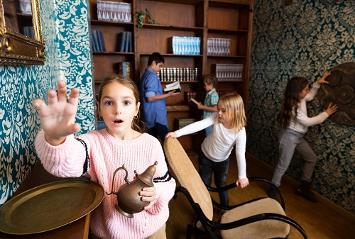Zdjęcie dzieci w wieku wczesnoszkolnym podczas gry fabularnej w stylizowanym pokoju: na pierwszym planie dziewczynka ze zdziwioną miną, ubrana na różowo wyciągająca przed siebie jedną rękę, drugą trzyma mosiężny imbryk, w tle dwóch chłopców przeglądających ksiązki przy regale, dwie dziewczynki, fotel bujany, na ścianach turkusowo tapety z białymi motywami kwiatowymi, i obrazy