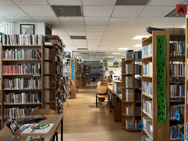 Korytarz w bibliotece. Po dwóch stronach stoją regały pełne książek. Na jednym z regałów naklejony jest zielony plakat z pionowym napisem  "lit obyczajowa"