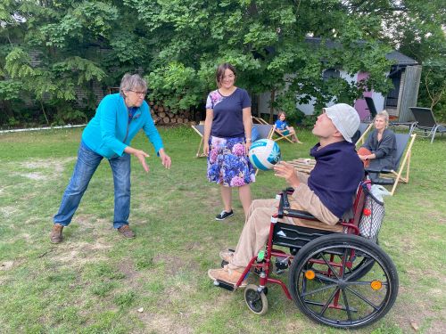 Trzy osoby grają na świeżym powietrzu w piłkę, rzucając ją do siebie, jeden z graczy siedzi na wózku, w tle widać kobietę, która siedzi na leżaku i obserwuje grę