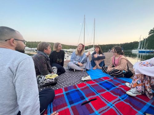 Grupa ludzi siedzących na kocach w kręgu na kei, na pierwszym planie jaskrawy koc w czerwoną i szafirową kratę, w tle jezioro i wystające maszty zacumowanych żaglówek