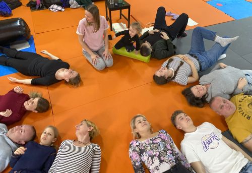Kilkanaście osób - dorosłych i dzieci - leży w kręgu na pomarańczowej macie, leżą na plecach z głowami skierowanymi do środka kręgu, wykonując ćwiczenia relaksacyjne