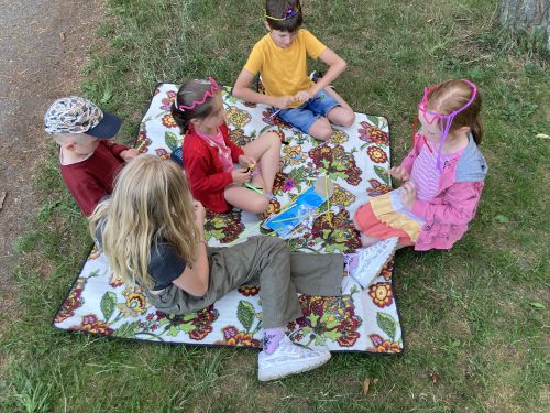 Dwóch chłopców i trzy dziewczynki ubrane w kolorowe letnie stroje siedzi na wzorzystym kocu piknikowym rozłożonym na trawie, część z nich ma na głowach opaski z drucików kreatywnych, wszyscy tworzą ozdoby z kolorowych drucików kreatywnych