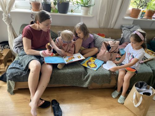 Zdjęcie wykonane w domu, na szarej kanapie siedzi młoda kobieta i troje małych dzieci, kobieta czyta dziecięcą książkę, na rękę ma nałożoną pacynkę