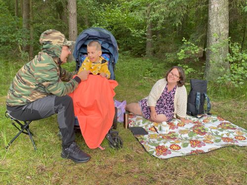 Zdjęcie pikniku rodzinnego, w którym uczestniczy dwoje dorosłych i dziecko, na trawie na rozłożonym kolorowym kocu siedzi kobieta, na stołku campingowym siedzi mężczyzna, pomiędzy nimi chłopiec w wózku, przykryty czerwonym kocem, wszyscy rozmawiają i uśmiechają się, w tle zieleń lasu