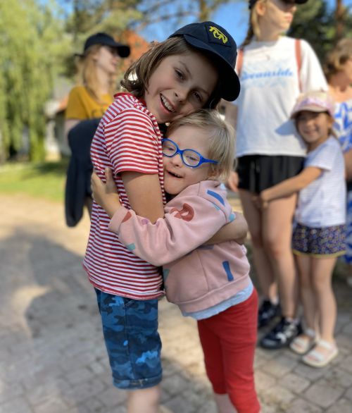 Zdjęcie dwójki dzieci, które uśmiechają się i przytulają do siebie, w tle inne osoby oraz zieleń, zdjęcie wykonane w porze letniej