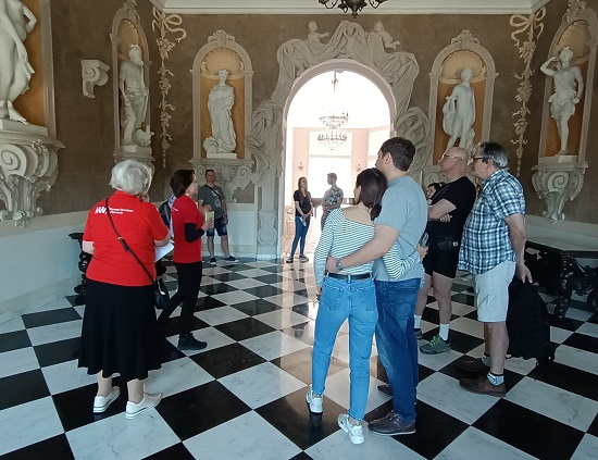 Zdjęcie wykonane w Muzeum Wnętrz w Otwocku: wolontariuszki w czerwonych koszulkach wolontariatu muzealnego, zwiedzający, na ścianach rzeźby, podłoga w biało-czarną szachownicę