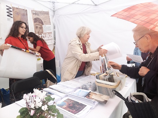 Zdjęcie wolontariuszek pomagających na stoisku MNW podczas Warszawskich Dni Seniora, trzy wolontariuszki w czerwonych koszulkach stoją przy stoliku, na stoliku są umieszczone wydruki kolorowe, plakaty, po drugiej stronie stolika seniorzy rozmawiający z wlontariuszkami