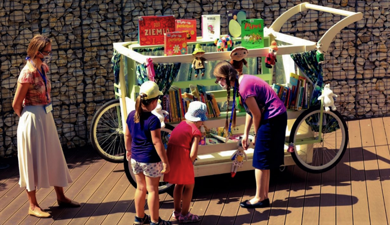 Wózek wystawienniczy z książkami dla dzieci obsługiwany przez wolontariuszkę