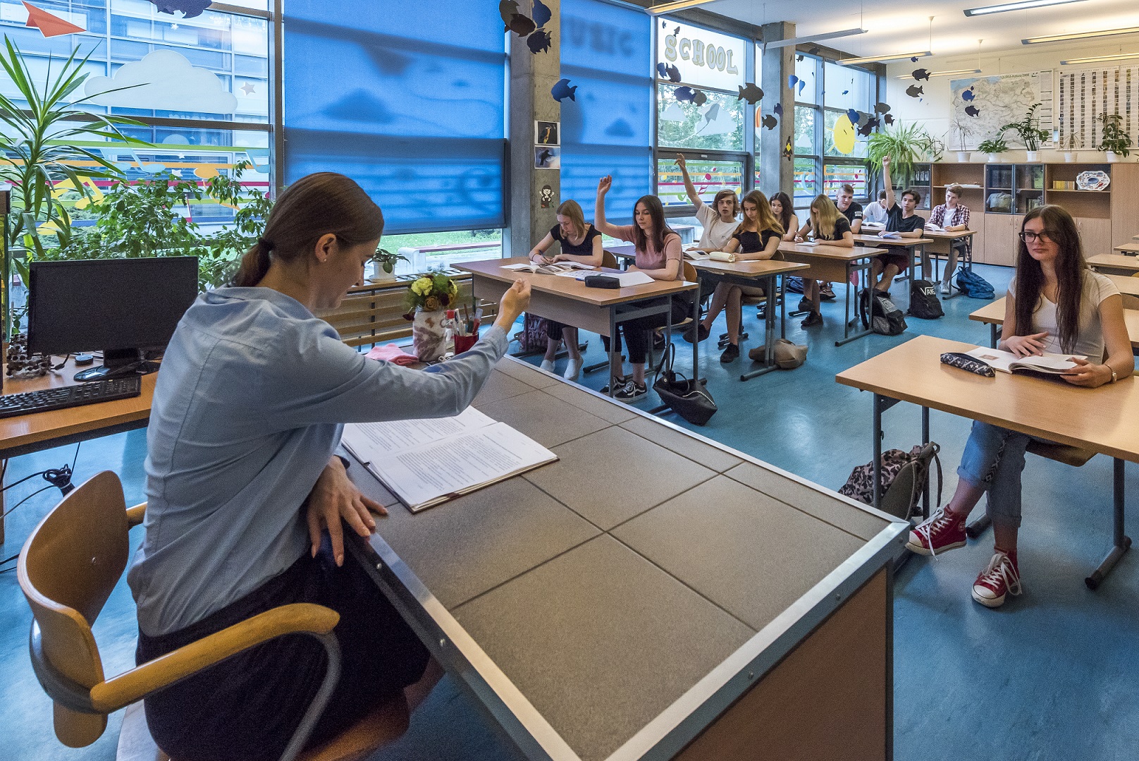 nauczycielka z uniesioną ręką przy biurku frontem do uczniów w ławkach 