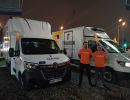 Dwóch wolontariuszy w pomarańczowych bluzkach swoi przed furgonetkami do transportu