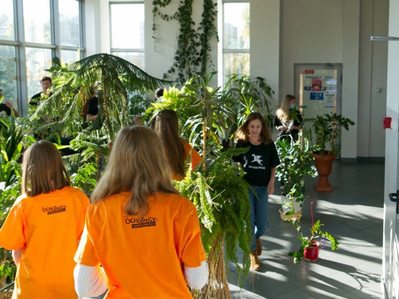 Wolontariuszki odwrócone plecami w koszulkach z logo projektu Ochotnicy warszawscy, w tle osoby i rośliny w doniczkach.