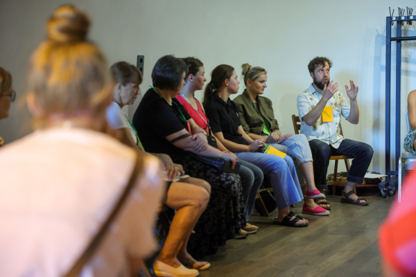 Wolontariusze i wolontariuszki siedzą w okręgu, Na środku znajduje się prowadzący warsztaty ze storytellingu, który tłumaczy coś reszcie grupy.