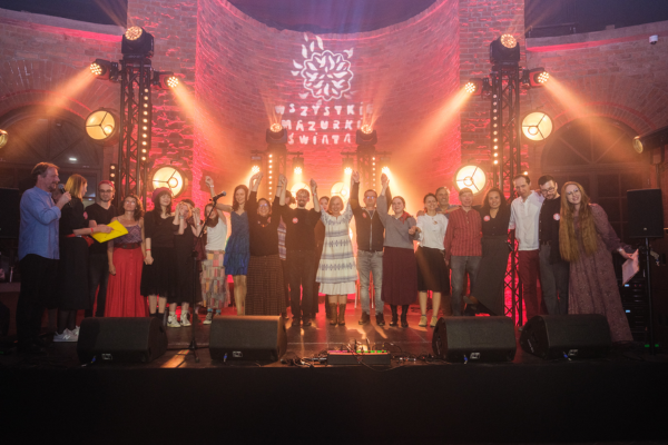 Muzykanci i organizatorzy na scenie podczas Festiwalu, dziękują za występ. Za nimi reflektory oraz mur z oświetlonej na czerwono cegły.