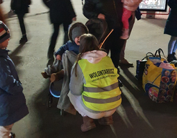 Zmrok. Wolontariuszka kuca przed dzieckiem w wózku, które płacze. W tle ludzie.