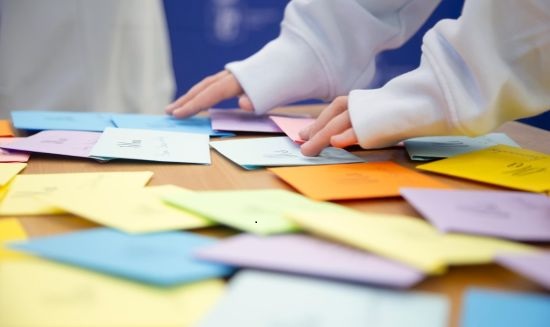 Kolorowe koperty rozłożone na stole, dwie dłonie, które sięgają po koperty