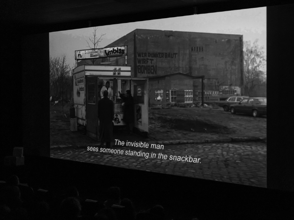 Ekran kinowy podczas festiwalu Let's Doc wyświetla czarno-białą klatkę filmu, na której widać dwie osoby stojące przy przyczepie z przekąskami