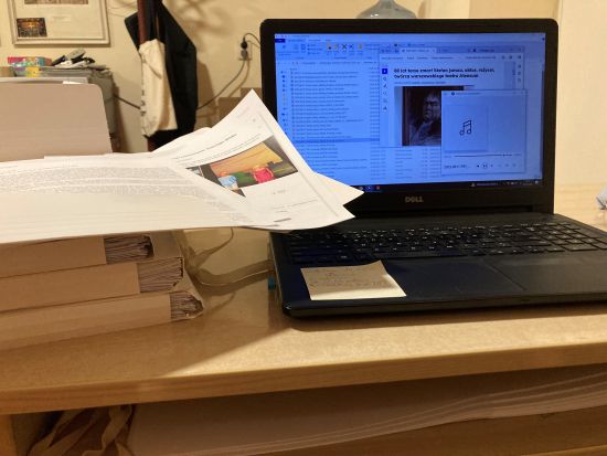 Laptop, stojący na biurku z otwartym plikiem głosowym, obok laptopa, po lewej stronie, stos materiałów drukowanych