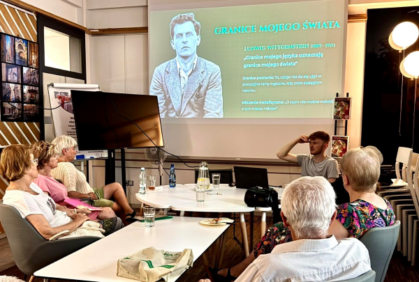 Grupa osób siedzi wokół stołu. Przy prawej krawędzi stołu siedzi Michał Kudliński. Za nim wyświetla się slajd z prezentacji "Granice mojego świata. Ludwig Wittgenstein"