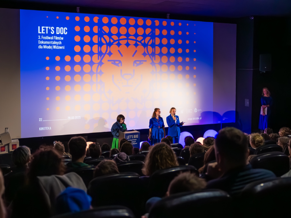 Przemówienie organizatorek festiwalu Let's Doc na sali kinowej, która zapełniona jest młodymi widzami i widzkami.