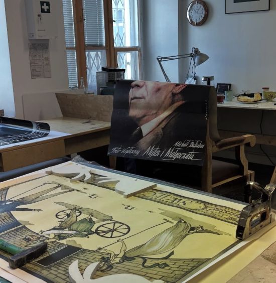 Pokój wypełniony archiwalnymi afiszami teatralnymi, rozmieszczonymi w różnych miejscach: na stole, na oparciu fotela, na biurku, w tle zegar