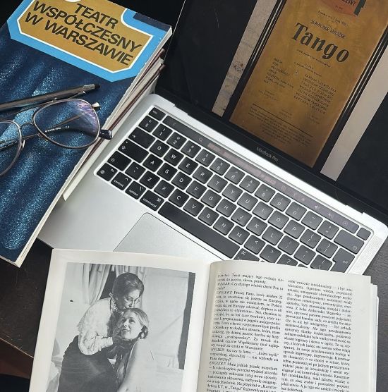 Po lewej stronie stos książek, z których górna ma tytuł: "Teatr Współczesny w Warszawie", na książce lezą okulary, po lewej stronie otwarty laptop z otwartym afiszem dotyczącym spektaklu "Tango" Sławomira Mrożka, na pierwszym planie otwarta książka z czarnobiałym zdjęciem dwóch kobiet