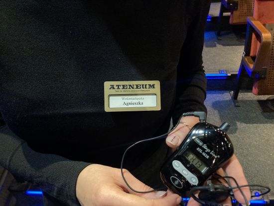 Aparat do audiodeskrypcji, trzymany w rękach przez wolontariuszkę, wolontariuszka ubrana na czarno, z przypiętym do bluzki złotym identyfikatorem "Teatr ATENEUM wolontariuszka Agnieszka"