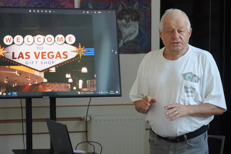 Pan Tadeusz stoi na tle obrazów i opowiada historię. Ubrany w białą koszulkę i jeansy. W tle na ekranie wyświetlony jest napis "Welcome to Las Vegas Gift Shop"