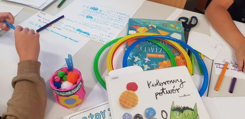 Na stole leżą książki i artykuły papiernicze oraz kartki z notatkami zrobionymi przez dzieci uczestniczące w warsztacie.