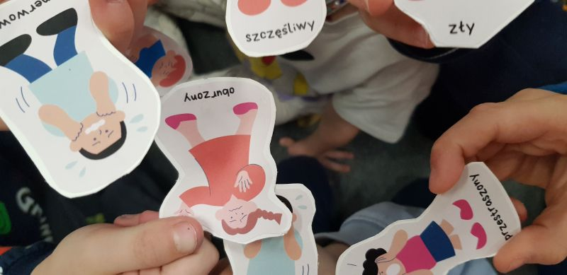 Grupa dzieci trzyma w rękach naklejki przedstawiające postaci, które reprezentują różne emocje.