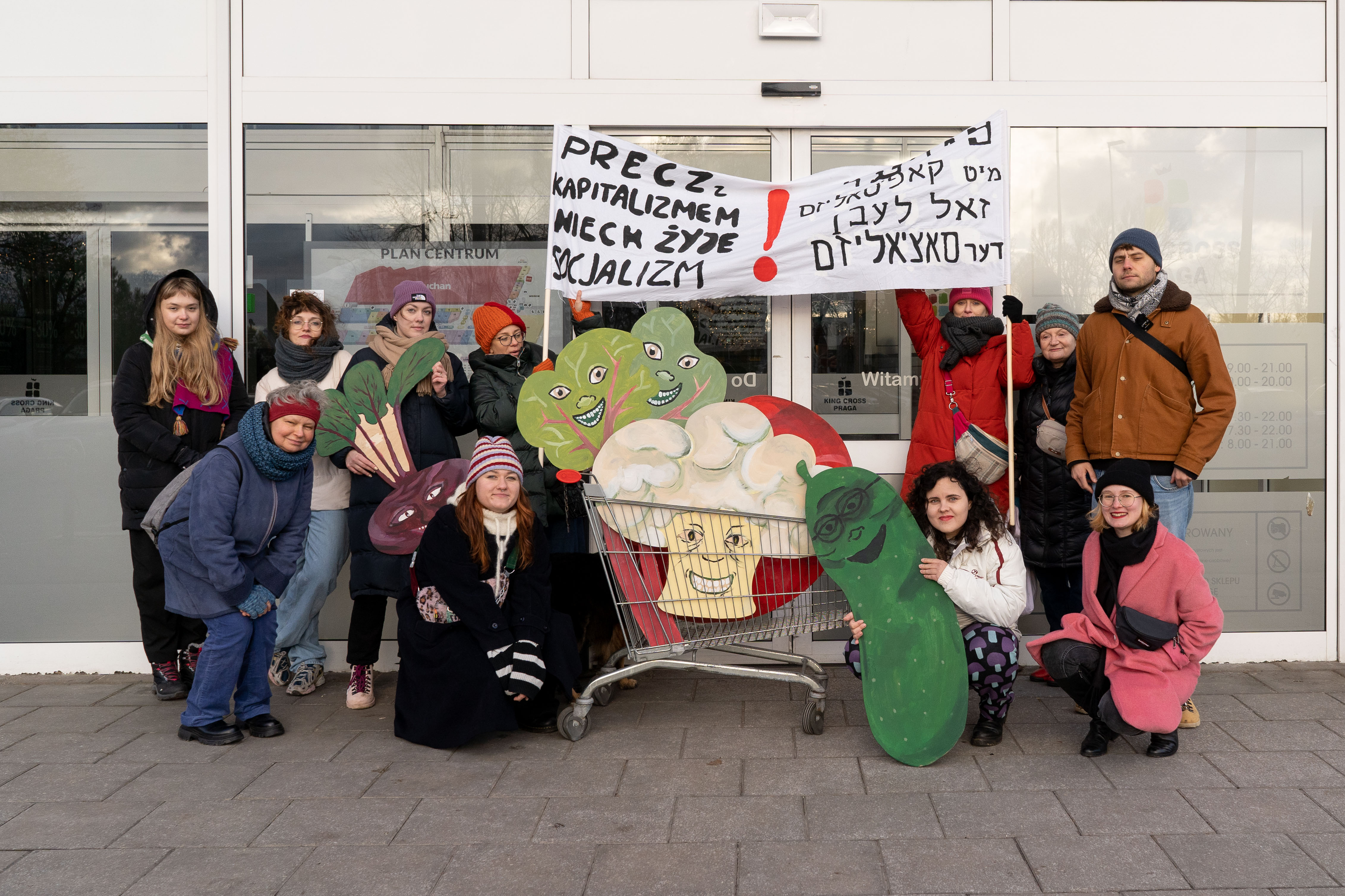 Grupa ludzi stojąca z wózkiem zakupowym przed supermarketem z papierowymi warzywami