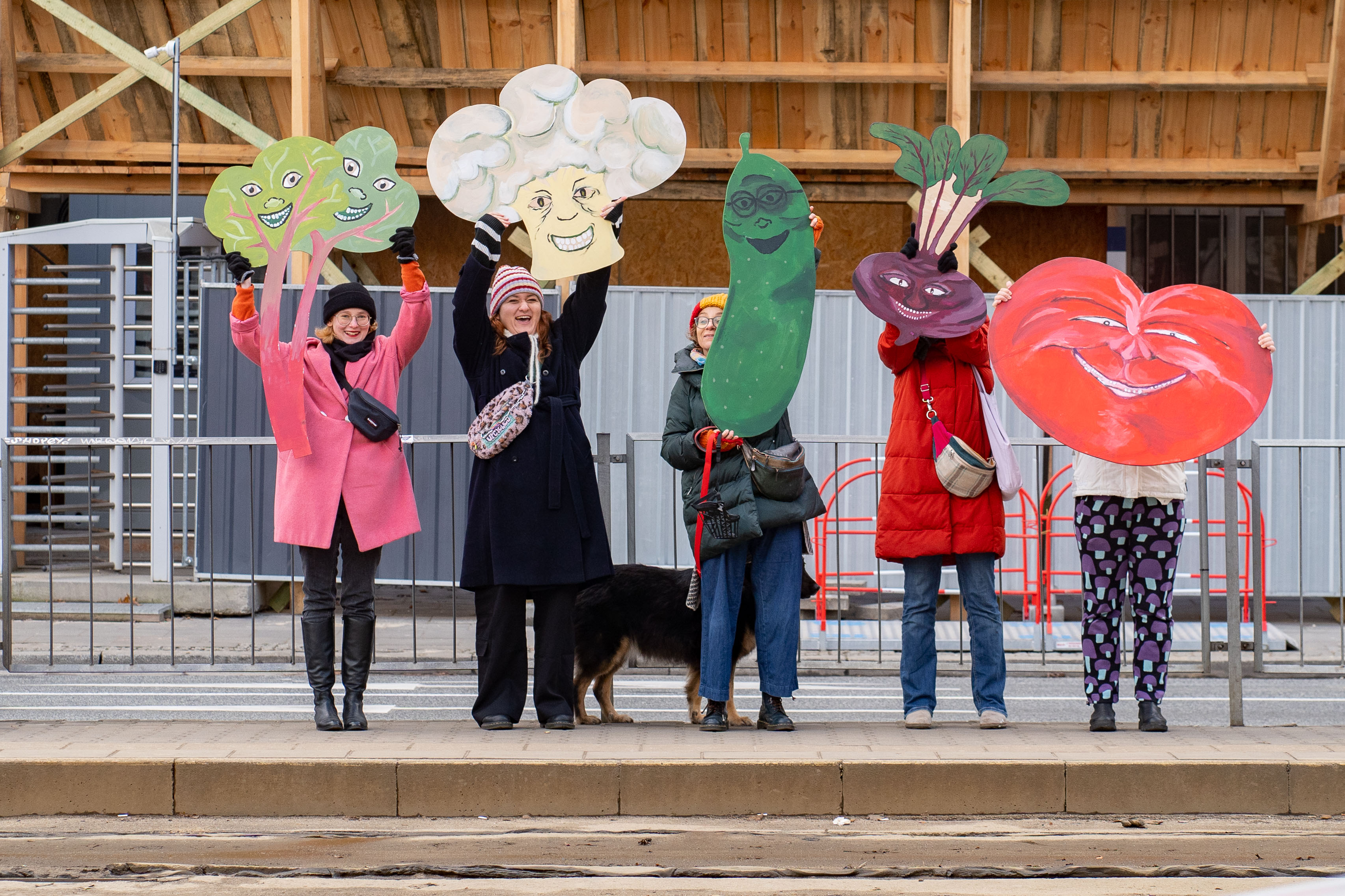 Grupa osób stojąca na przystanku i trzymająca duże warzywa wycięte z papieru.