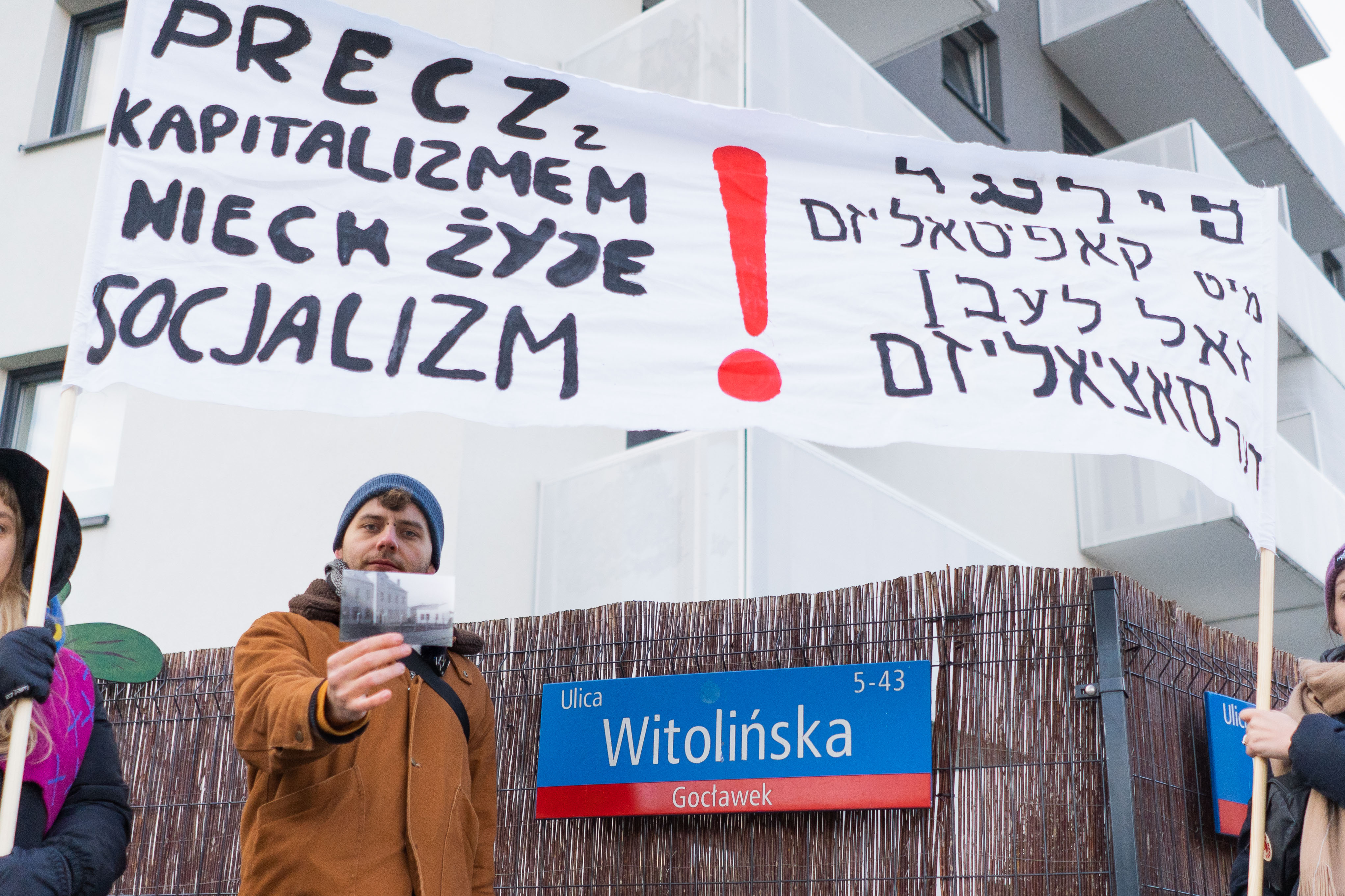 Mężczyzna stojący przy ulicy Wierzbickiej i trzymający czarno-białe zdjęcie. W tle widać biały plakat z czarnym napisem "precz z kapitalizmem, niech żyje socjalizm"