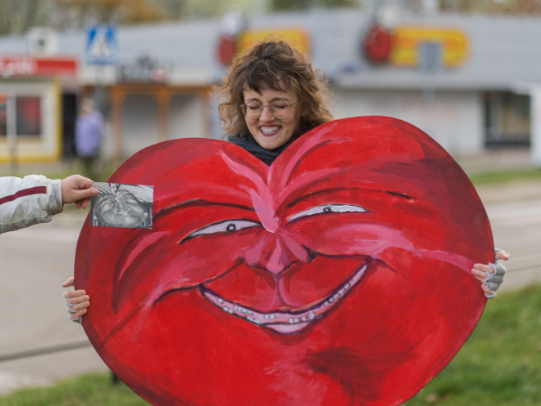 Usmiechnięta kobieta z kręconymi włosami i w okularach trzymająca duże serwone uśmiechnięte serce.