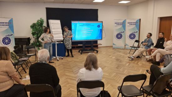 Zdjęcie z warsztatów prowadzonych w sali warsztatowej, uczestnicy siedzą na krzeslach ustawionych w kręgu, na dalszym planie dwie prowadzące, stojące przy flipcharcie i wyświetlonej prezentacji