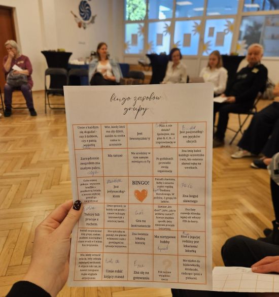 Na pierwszym planie kartka z grą bingo, w tle sala warsztatowa i uczestnicy warsztatu siedzący w kręgu na krzeslach