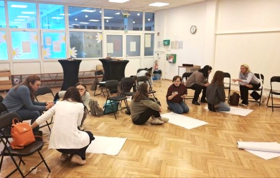 Zdjęcie wykonane w sali: na parkiecie są rozłożone duże arkusze białego papieru, przy których siedząc na podłodze lub na krzesłach w grupach po kilka osób dyskutują i pracują uczestnicy warsztatów  